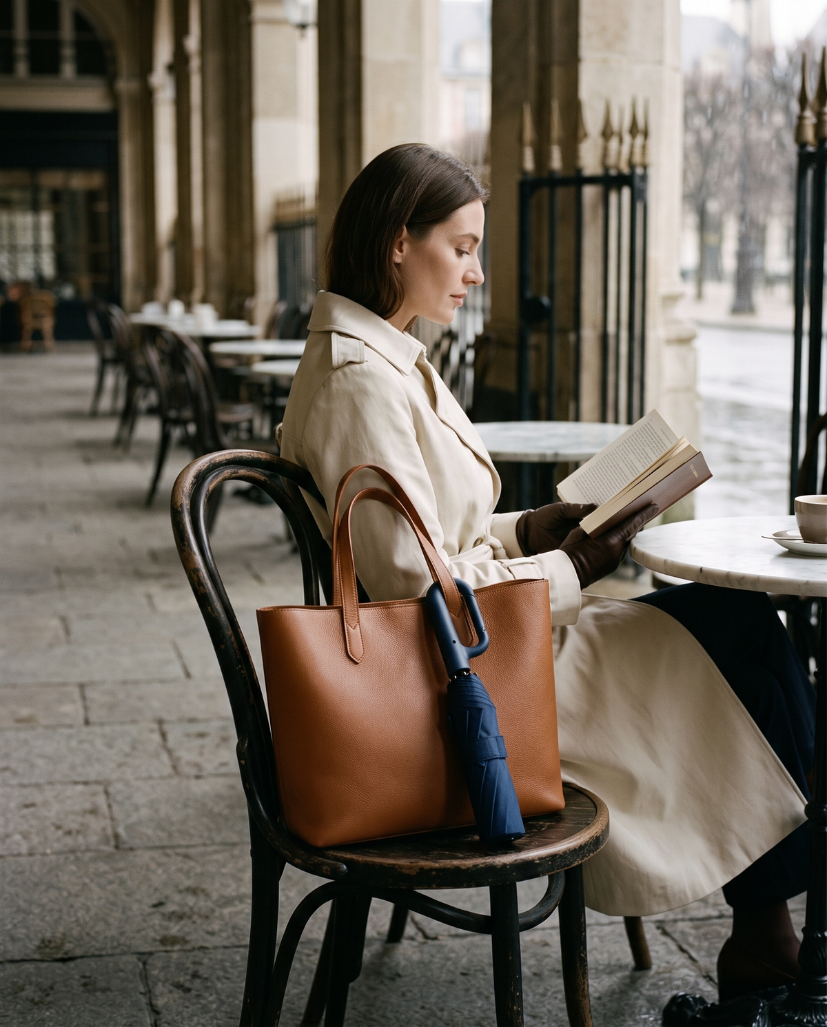 Woman reading at a café table in the rain, the navy Ombriel Loop hanging from the handle of her tan leather tote