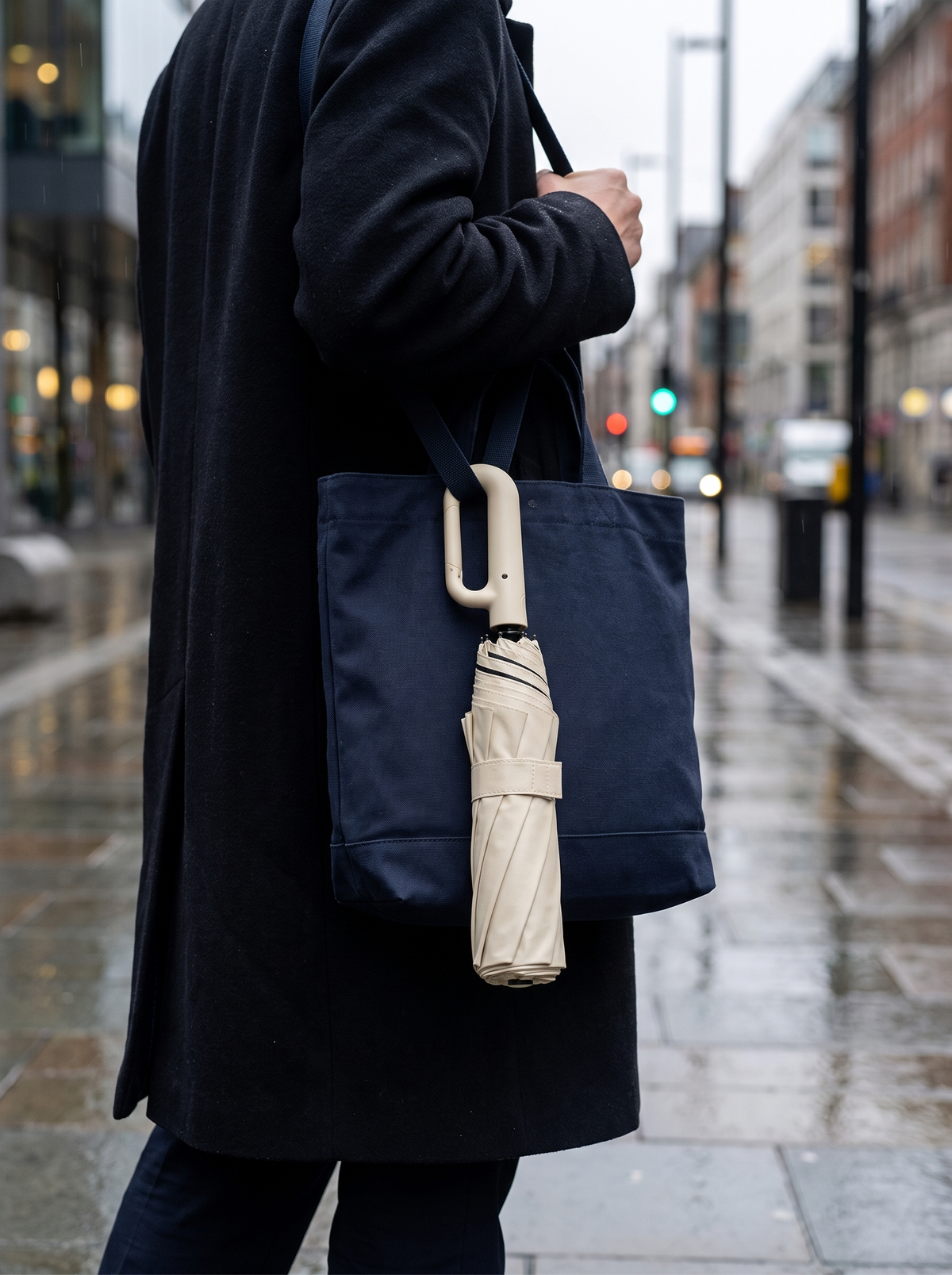 Ombriel Loop umbrella in beige hanging by its ring handle from the strap of a navy tote bag on a rainy city street