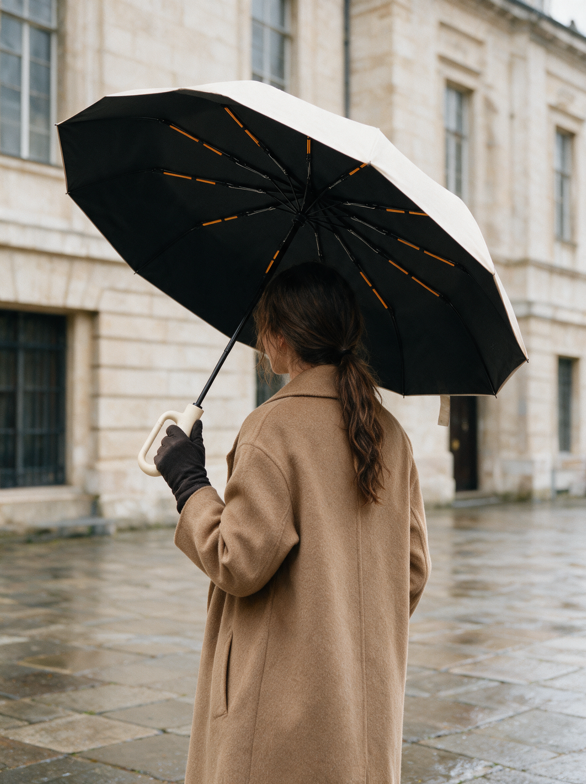Woman in a camel coat on a wet stone square holding the open beige Ombriel Loop tilted to show the black underside with orange fibreglass strips